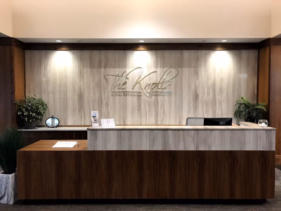 Reception desk in a senior living lobby with a wall sign reading "The Knolls," plants, and overhead lighting.