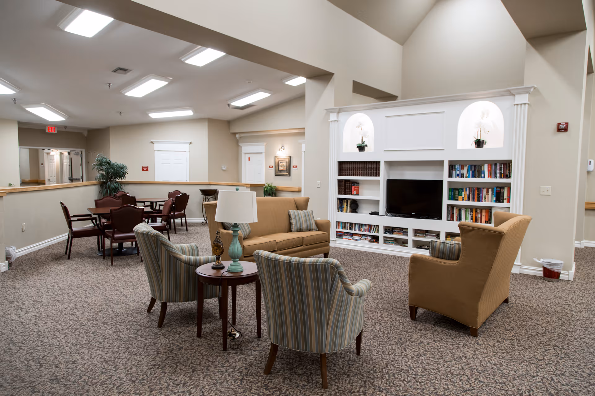 Spacious senior living common room with sofas and armchairs arranged around a TV and built-in bookshelves, and dining tables in the background.