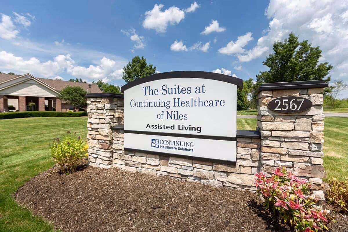 Stone entrance sign reading "The Suites at Continuing Healthcare of Niles Assisted Living" on a landscaped lawn with the facility building in the background.