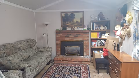 A cozy living room with a patterned sofa on the left, a brick fireplace with a painting above it in the center, and a wooden bookshelf filled with books and decorative items on the right. There is a floor lamp next to the sofa, a patterned rug on the tiled floor, and a wooden dresser with a small stool in front of it. Several hats hang on the wall near a window with floral curtains.