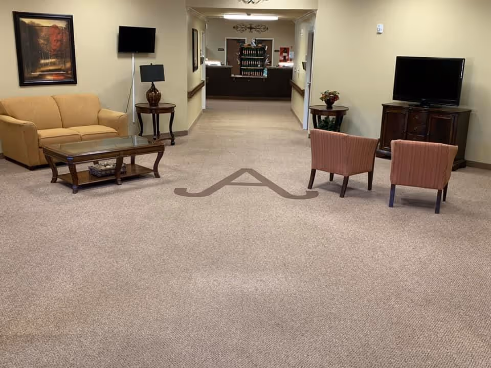 A spacious living room area in a healthcare and rehabilitation center featuring a beige sofa, two pink armchairs, a glass coffee table, two side tables with lamps and a flower vase, a wall-mounted TV, and a large flat-screen TV on a wooden cabinet. The carpet has a large letter 'A' design in the center. The room leads to a reception or nurse station area in the background.