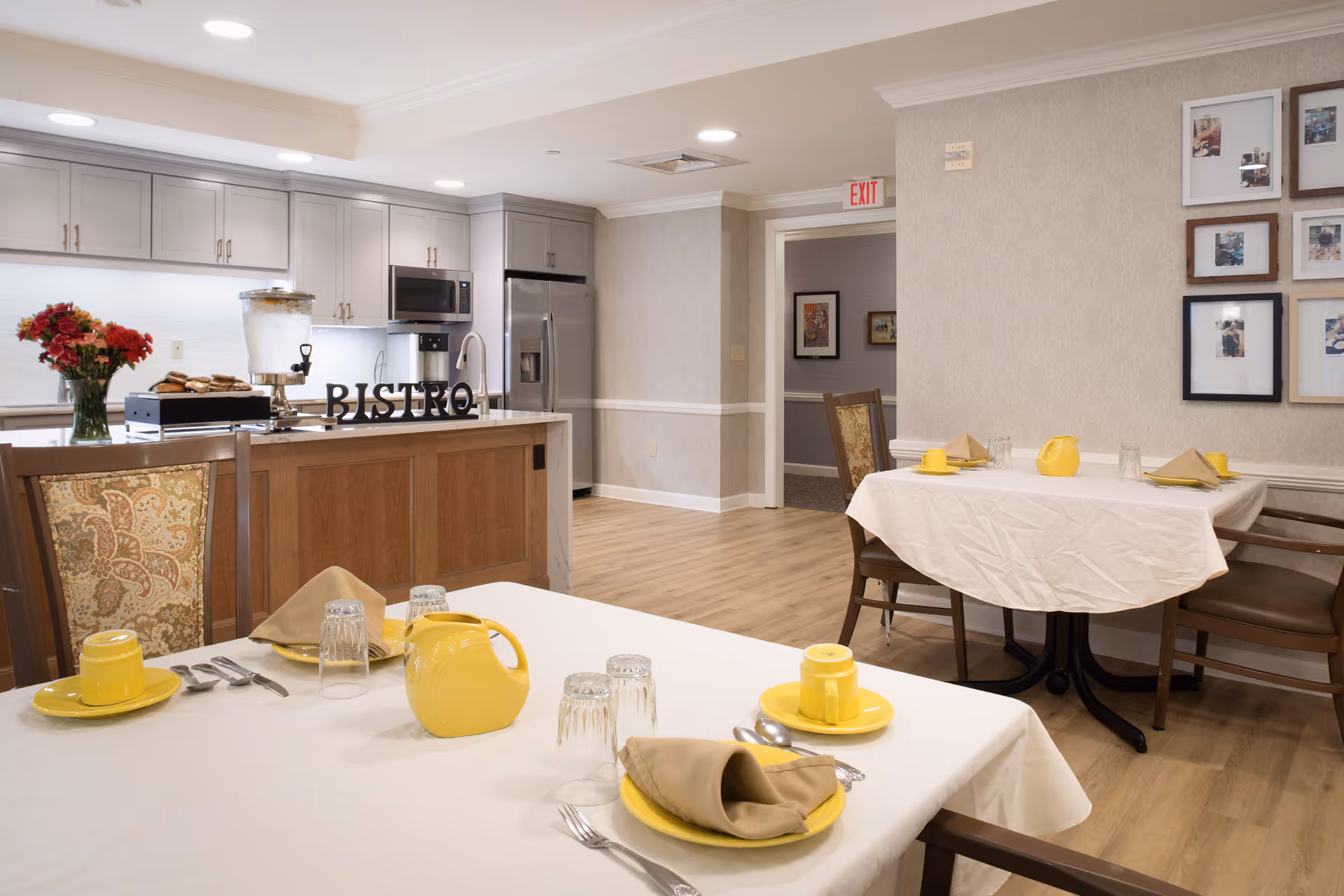 A dining area in a senior living facility featuring two tables set with yellow cups, saucers, plates, beige napkins, and glasses. The room has light wood flooring and beige patterned wallpaper. In the background, there is a kitchen area with gray cabinets, a stainless steel refrigerator, microwave, and a countertop with a water dispenser and a tray of cookies. The word 'BISTRO' is displayed on the kitchen counter. Several framed photos hang on the wall to the right.