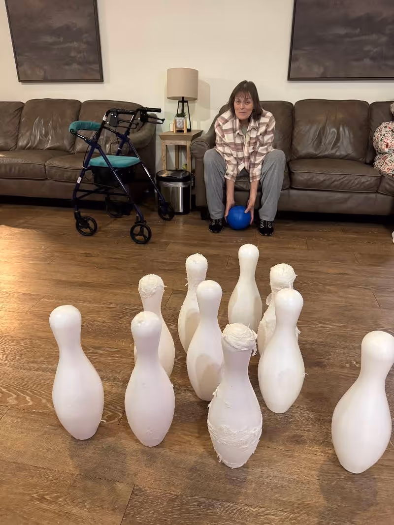 A woman sitting on a brown leather couch indoors, holding a blue ball and preparing to roll it towards a set of white bowling pins arranged on a wooden floor. A walker is positioned next to the couch, and there is a small side table with a lamp and a trash can behind her.