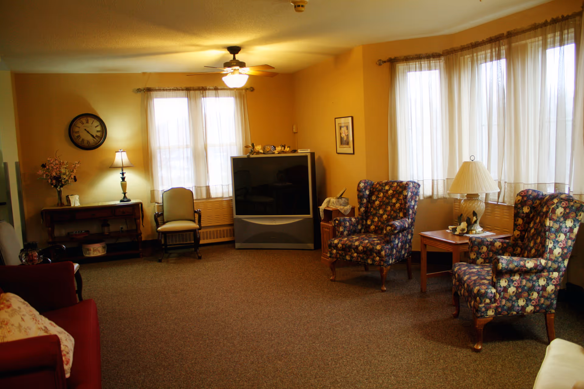 A cozy living room area with beige walls and carpeted floor. The room features two floral-patterned armchairs with a wooden side table and lamp between them, a large older-style television, a wooden chair, a small table with a lamp and flowers, and a wall clock. Light filters in through sheer curtains on multiple windows.