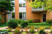 Brick-faced retirement community building exterior with ground-floor windows, a small balcony, a doorway, and landscaped flowerbeds and shrubs.