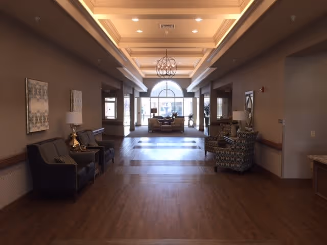 Spacious, well-lit lobby of a senior living facility with seating, chandeliers, and a large arched window at the far end.