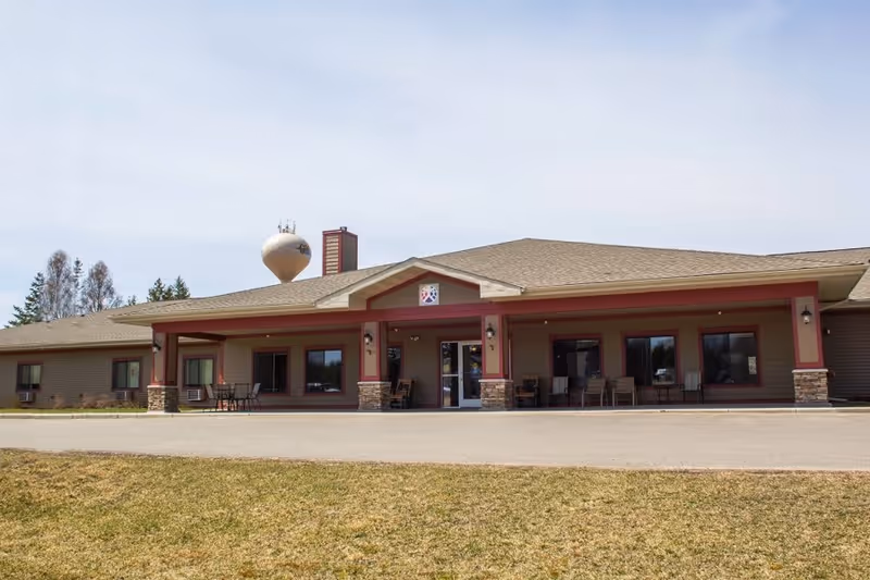 Exterior front view of a single-story senior living facility building with a covered entrance supported by stone pillars and red trim. There are several chairs and tables on the porch area. A water tower is visible in the background behind the building.