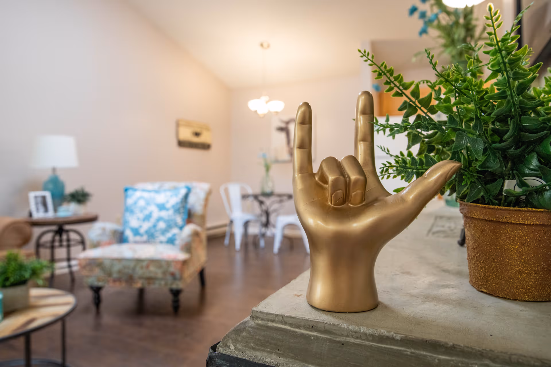 A cozy living room area with a floral armchair with a blue and white pillow, a side table with a lamp and small plant, and a dining table with white chairs in the background. In the foreground, there is a golden hand sculpture making the 'I love you' sign in American Sign Language, next to a potted green plant.