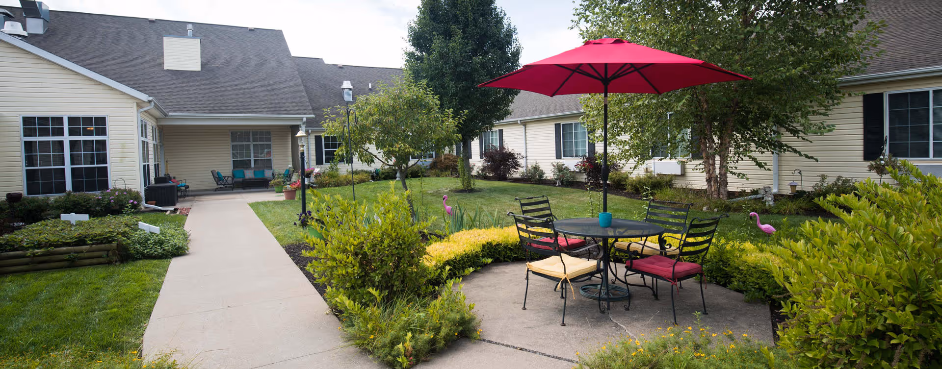Outdoor courtyard area at Bickford of Quincy featuring a round glass table with four chairs, two with red cushions and one with a yellow cushion, under a red umbrella. The courtyard is surrounded by green bushes, small trees, and beige buildings with windows and doors. A concrete pathway leads through the garden area.