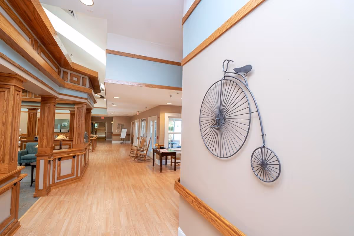 Interior hallway of a senior living facility with light wood flooring and wood trim. On the right wall, there is a decorative metal sculpture of a vintage penny-farthing bicycle. The hallway leads to a seating area with rocking chairs and tables, and there is a partial view of a lounge area with blue chairs on the left.