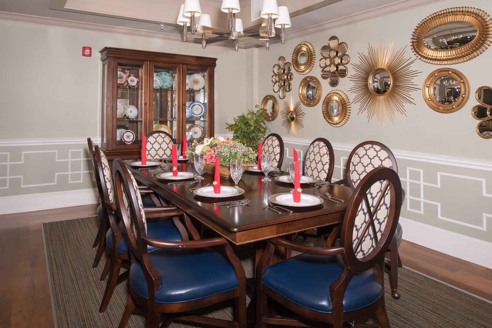 Formal dining room with a long wooden table set for eight, patterned chairs, a china cabinet, and decorative gold sunburst mirrors on the wall.