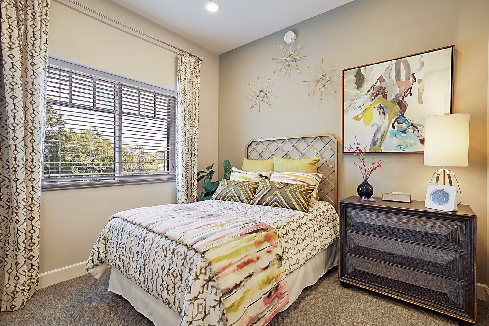 A cozy bedroom in a senior living facility featuring a bed with patterned bedding and multiple pillows, a wooden nightstand with a lamp, a clock, and a vase with flowers. The room has a large window with blinds and patterned curtains, abstract wall art, and decorative starburst wall sculptures.
