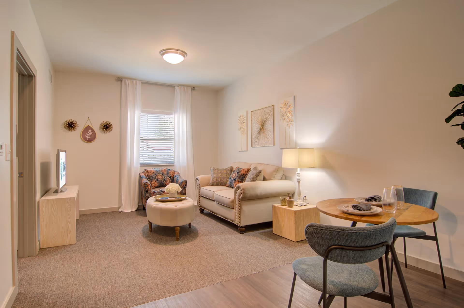 Cozy furnished living room with a sofa, patterned armchair, round ottoman, TV on a stand, floor lamp, and a small dining table for two.