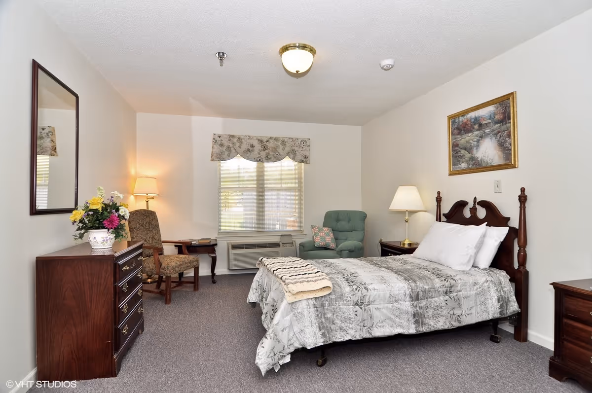 A senior living bedroom with a bed covered in a gray patterned comforter and white pillows. There is a wooden headboard, a nightstand with a lamp, a green armchair with a floral pillow, a wooden dresser with a vase of flowers, a mirror above the dresser, and a window with floral valance. The room has beige walls and gray carpet.