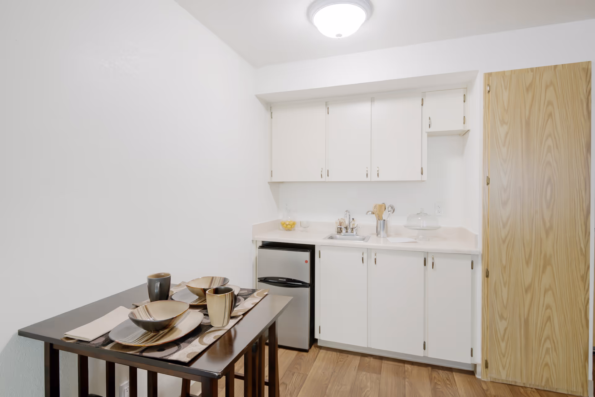 Small kitchenette with white cabinets, a mini fridge and sink, and a small dining table set with dishes.