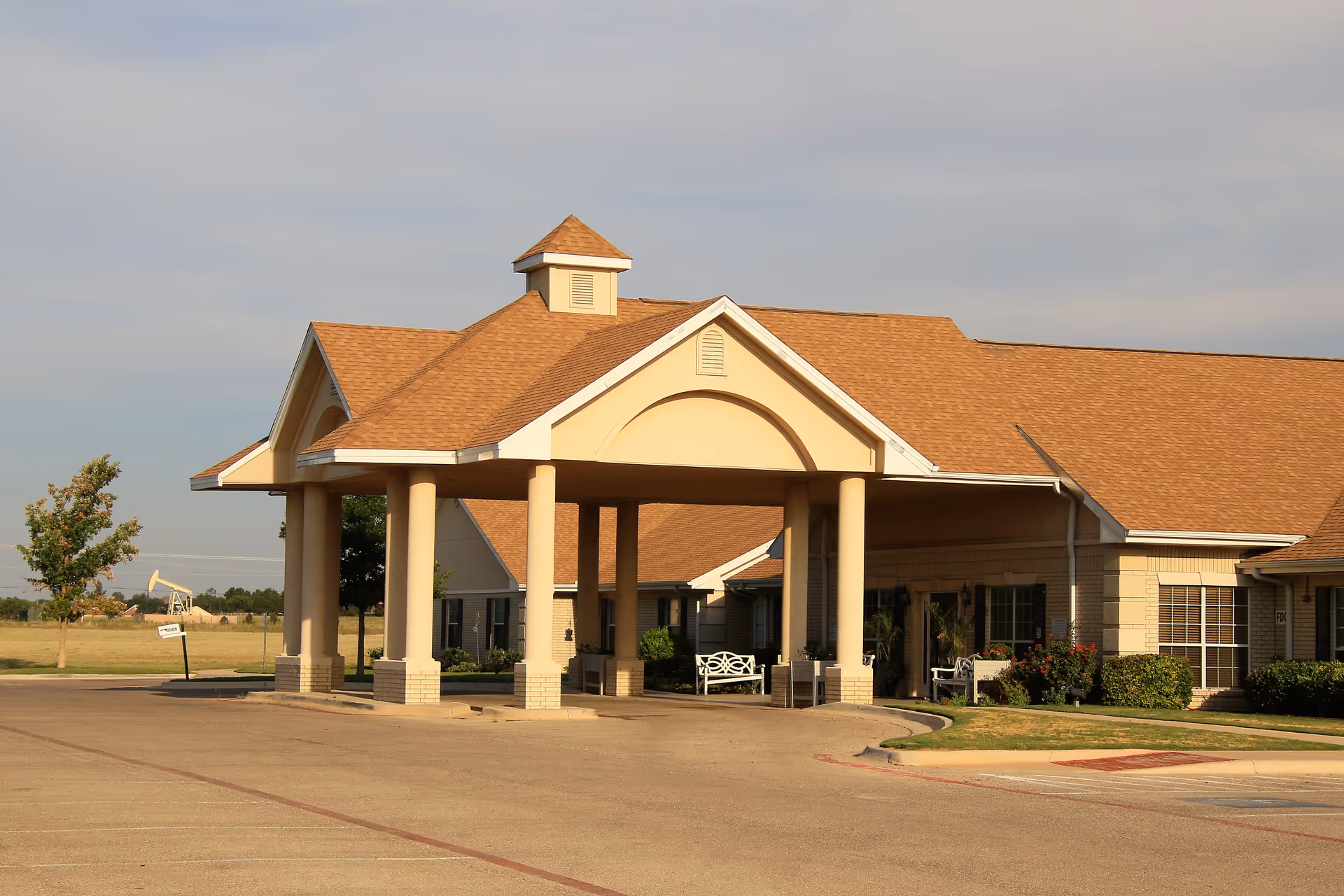 Exterior view of Midland Medical Lodge showing the front entrance with a covered drop-off area supported by columns, surrounded by a parking lot and landscaped greenery under a clear sky.