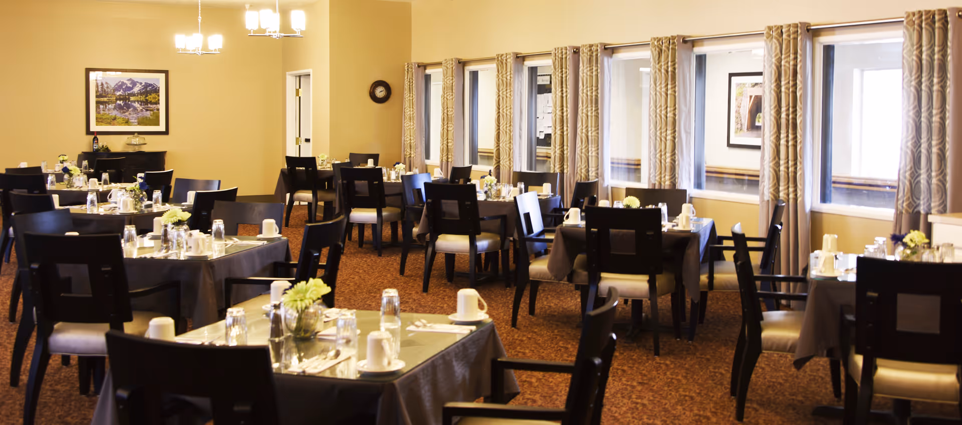 A dining room with multiple tables covered in dark tablecloths, each set with white cups, glasses, silverware, and small flower arrangements. The room has beige walls, patterned curtains on the windows, and framed artwork on the walls. The lighting is warm with ceiling lights and a chandelier.