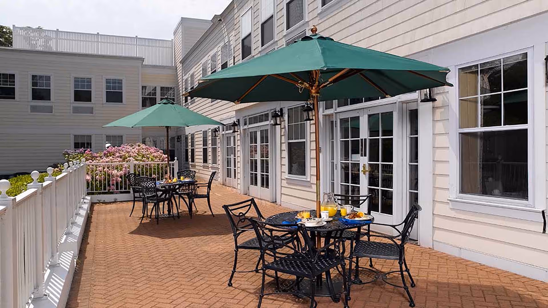 Outdoor patio area at Atria Bay Shore with black metal tables and chairs, green umbrellas providing shade, and a brick floor. The patio is adjacent to a light-colored building with multiple windows and glass doors. There are pink flowering bushes along the white railing on the left side.
