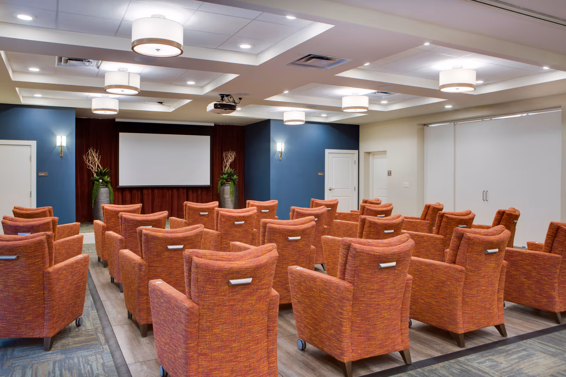 A well-lit conference or presentation room with multiple rows of orange upholstered chairs facing a large white projection screen. The room has a blue accent wall with two large decorative vases containing plants on either side of the screen. The ceiling features recessed lighting and modern circular light fixtures.