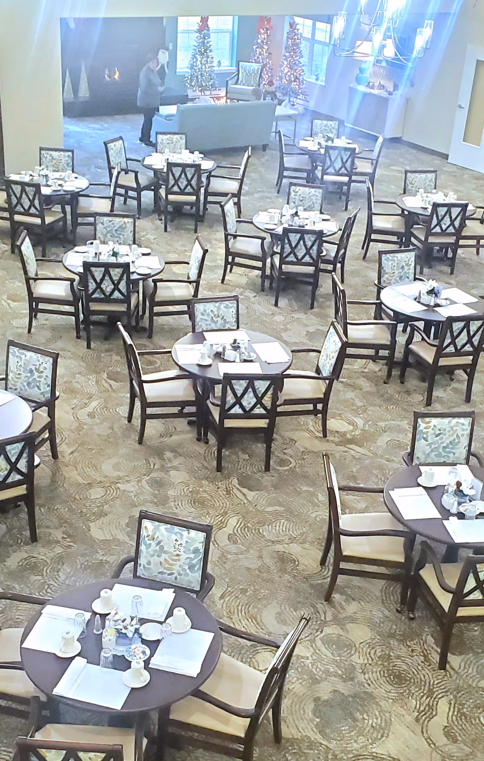 A spacious dining room with multiple round tables, each surrounded by four chairs with floral patterned backs. The tables are set with cups, glasses, and napkins. In the background, there is a seating area with a sofa and armchairs near a fireplace, decorated with two Christmas trees and holiday ornaments. A person is standing near the fireplace.