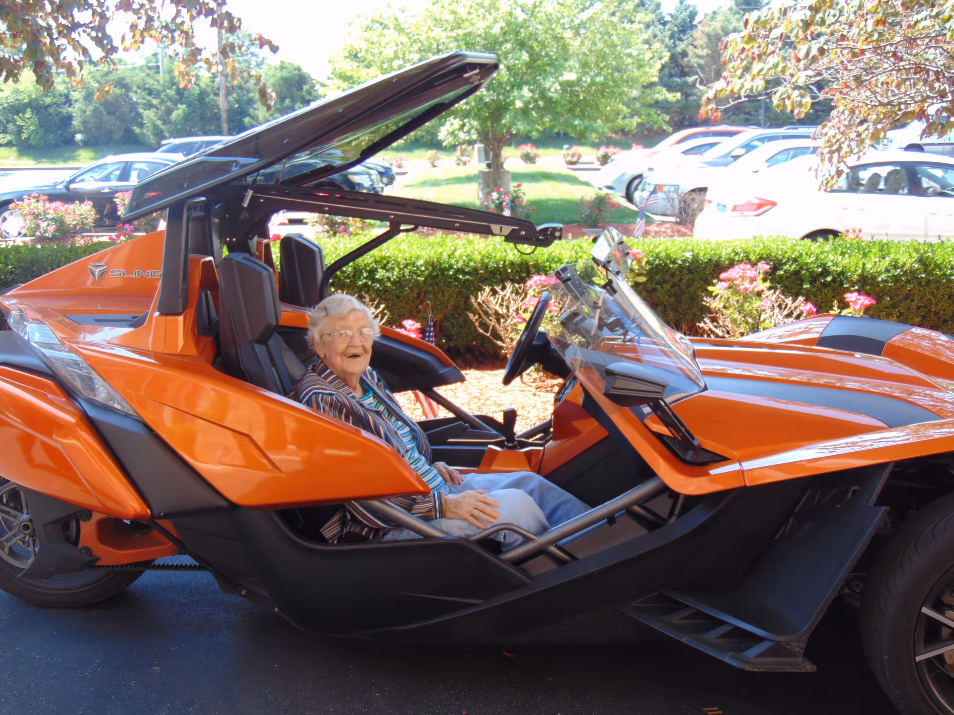 An elderly woman sitting and smiling inside an orange three-wheeled vehicle parked outdoors with cars and greenery in the background.