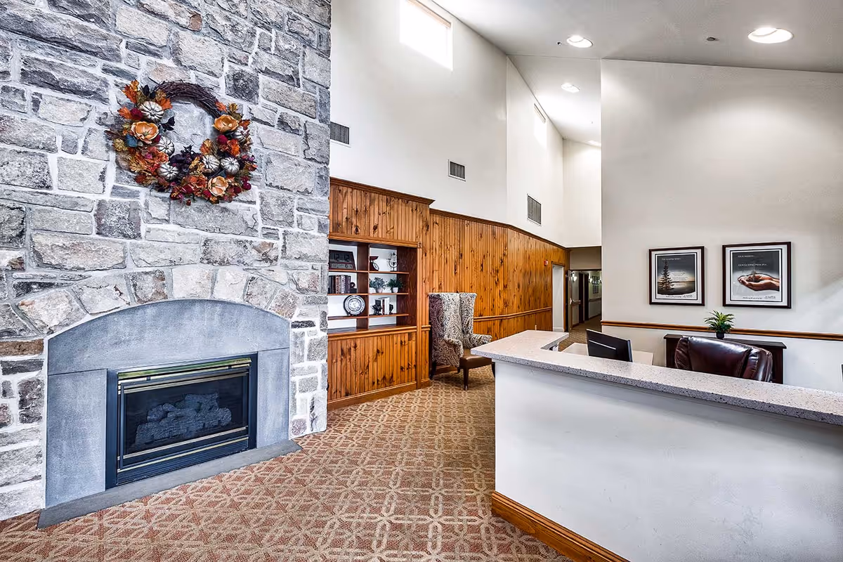 Interior view of a senior living facility reception area featuring a stone fireplace decorated with a fall-themed wreath, a wooden built-in bookshelf with decorative items, a patterned armchair, and a reception desk with a computer monitor and leather chair. The walls are white with wood paneling along the lower half, and two framed pictures hang on the wall behind the desk.