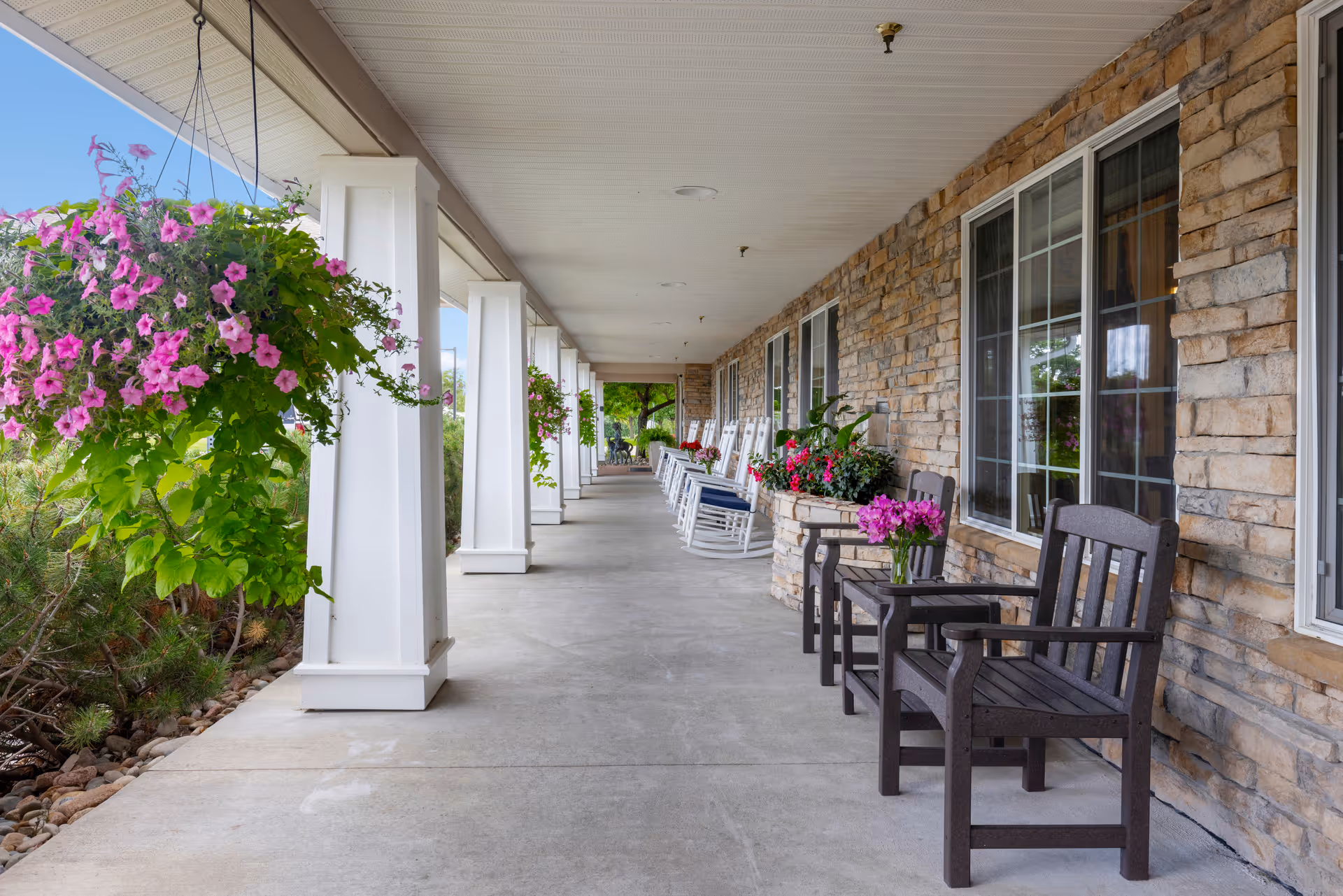 A long covered outdoor porch at Kipling Meadows Senior Living with hanging baskets of pink flowers, white columns, stone walls, large windows, and seating including dark wooden chairs with a small table and white rocking chairs further down the porch.