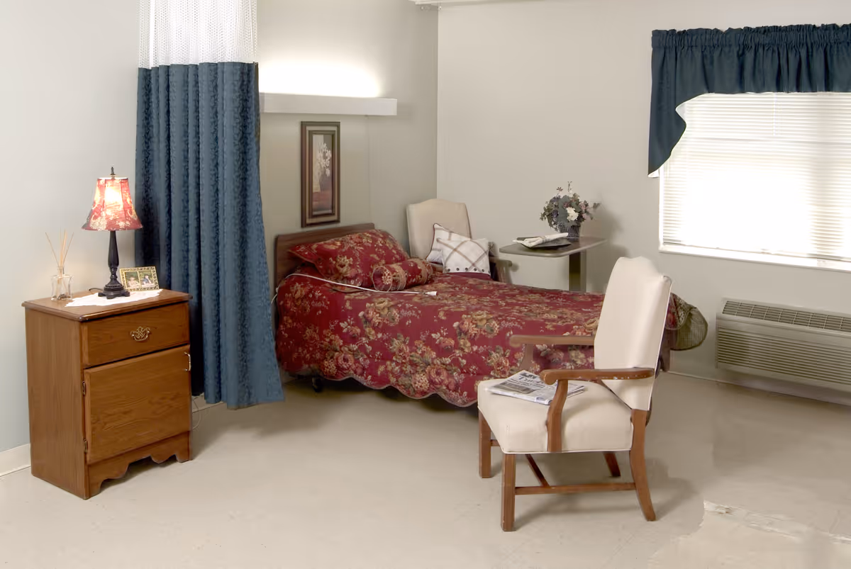 A tidy senior living bedroom with a single bed covered in a red floral bedspread, a wooden nightstand with a lamp, and an upholstered armchair.