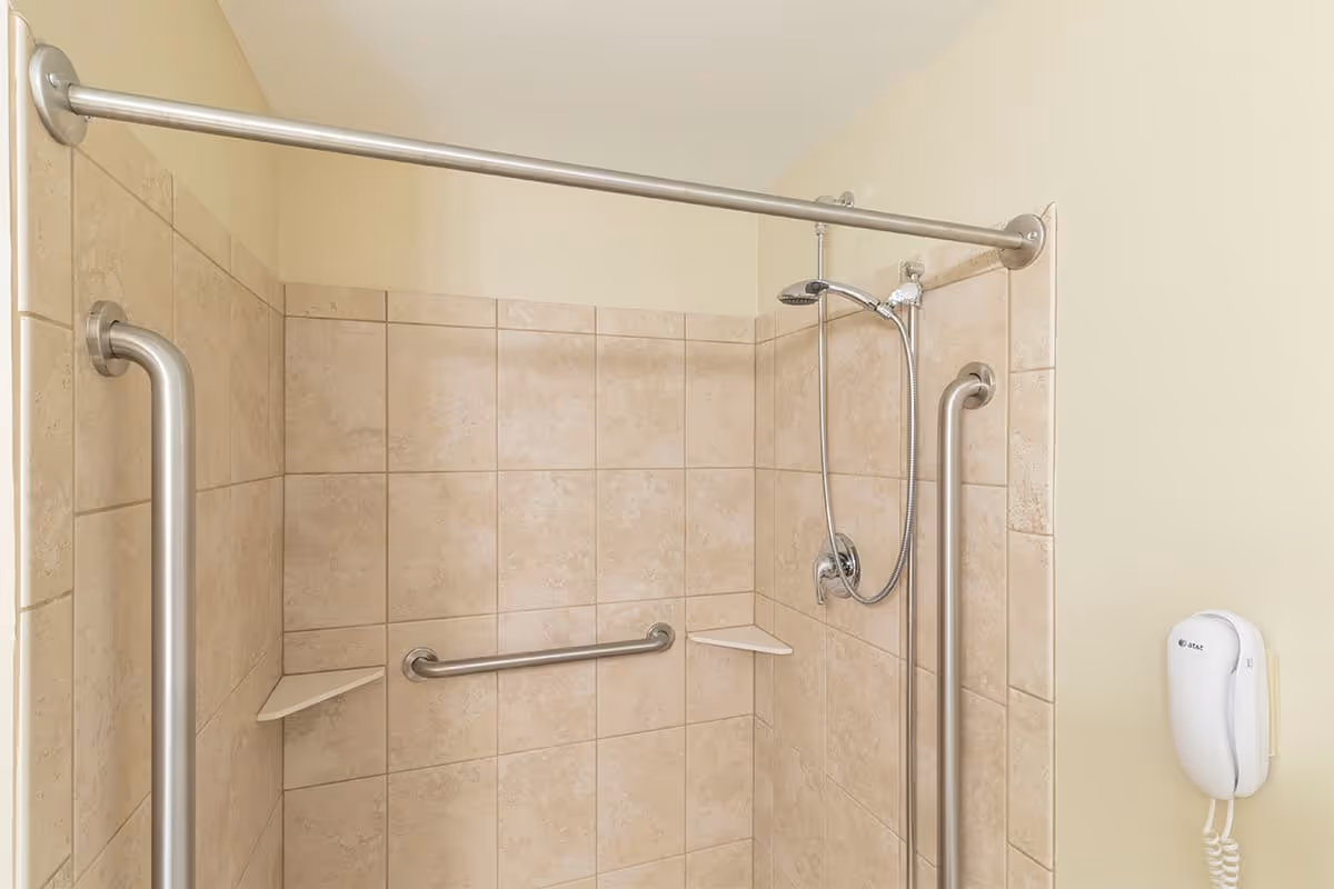 A tiled shower area with beige tiles, equipped with stainless steel grab bars on the side and back walls. There is a handheld showerhead attached to the wall and two small corner shelves. A wall-mounted white telephone is visible on the right side outside the shower area.