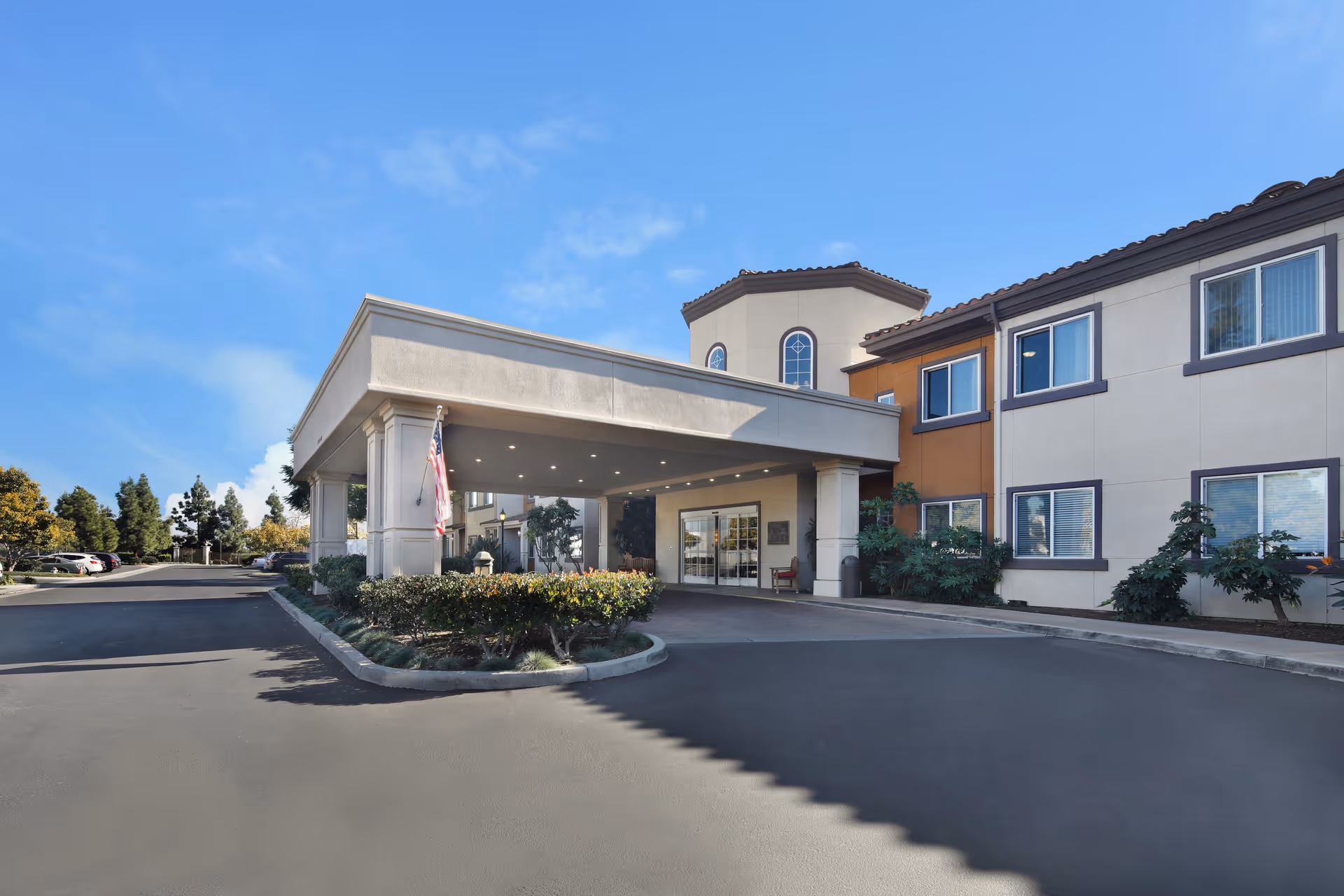 Exterior view of Ivy Park at Cerritos senior living facility entrance with a covered drop-off area, American flag, and surrounding landscaping under a clear blue sky.