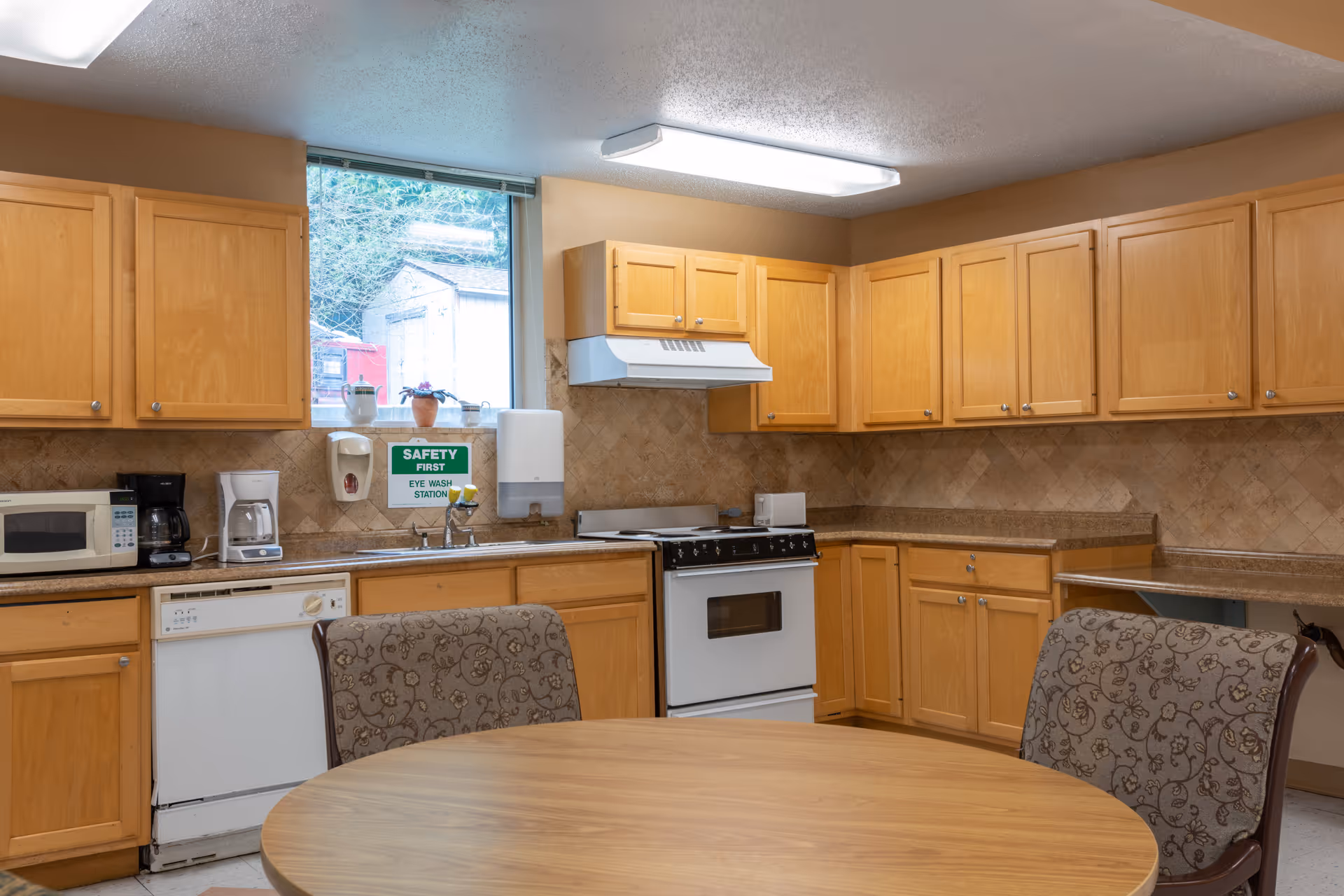 A communal kitchen with wooden cabinets, a round table and upholstered chairs, a stove, microwave, dishwasher and a sink beneath a window.