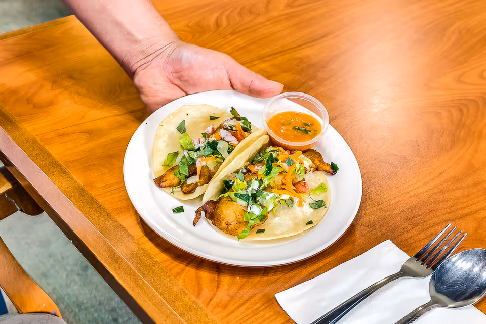 A hand holding a white plate with two fish tacos topped with lettuce, diced tomatoes, and herbs, accompanied by a small cup of orange dipping sauce on a wooden table. A fork and spoon rest on a white napkin beside the plate.