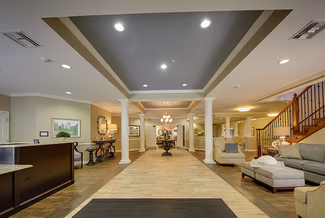Spacious and well-lit senior living facility lobby with a central walkway flanked by white columns. On the right side, there is a seating area with sofas, armchairs, and an ottoman. On the left side, there is a reception desk and decorative tables with lamps and artwork on the walls. A staircase with wooden handrails is visible on the right side.