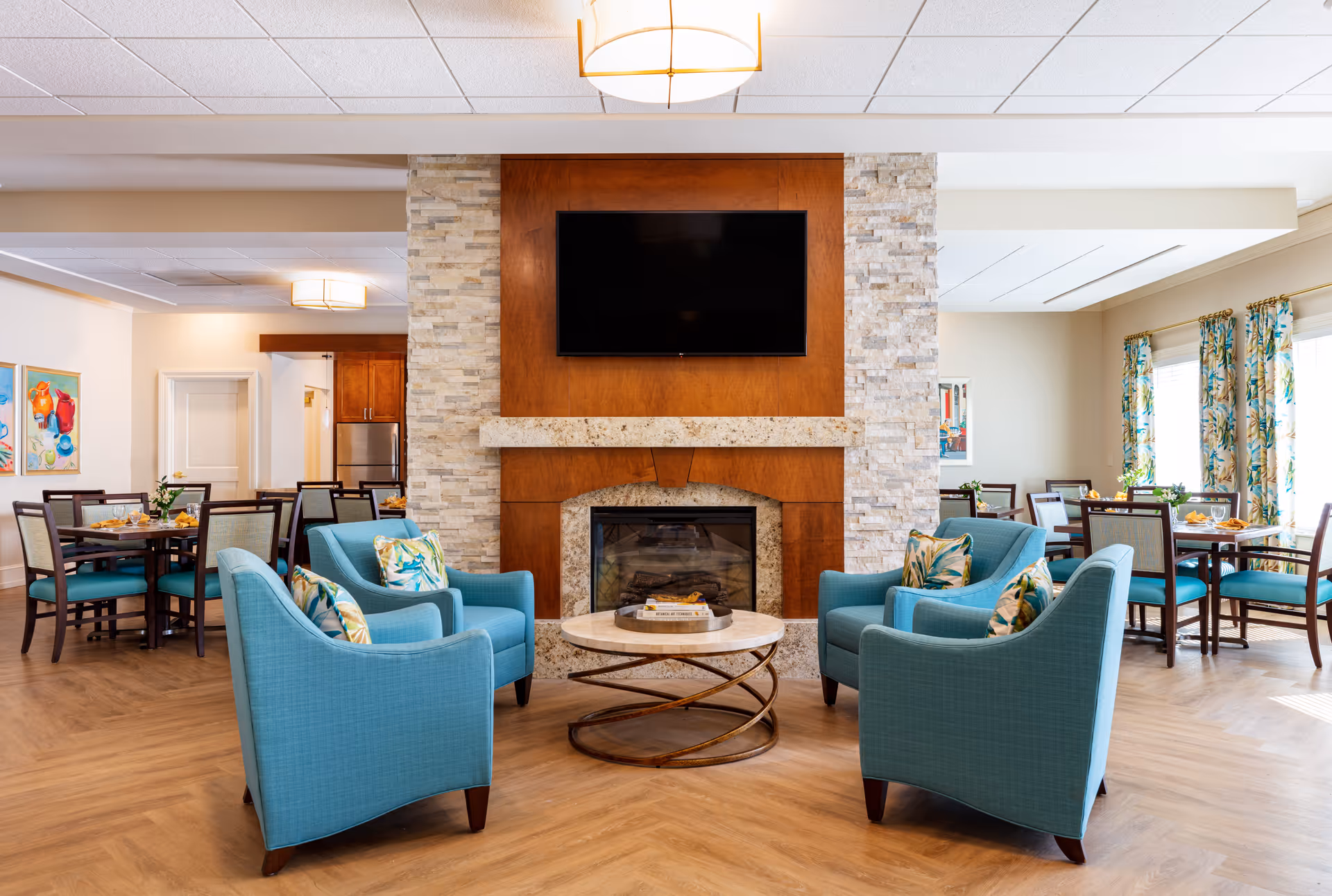 A cozy senior living facility common area with four teal armchairs arranged around a round coffee table in front of a stone and wood fireplace with a mounted flat-screen TV above it. Behind the seating area, there are dining tables with chairs, colorful artwork on the walls, and windows with floral curtains letting in natural light.