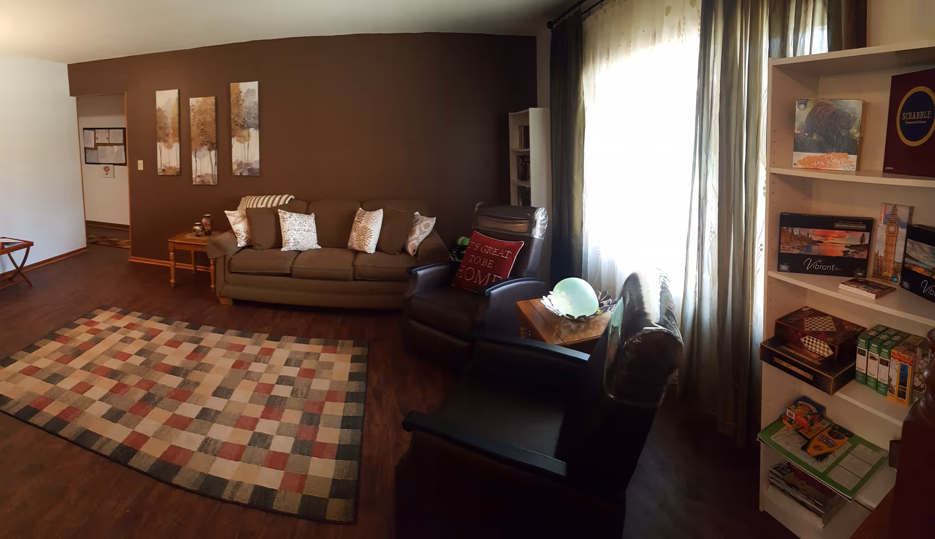A cozy living room with a brown sofa adorned with patterned pillows, two dark leather armchairs, a wooden side table with a glowing decorative orb, and a colorful checkered rug on a wooden floor. The room has a dark brown accent wall with three framed tree paintings and a white bookshelf filled with books and games near a window with sheer curtains.