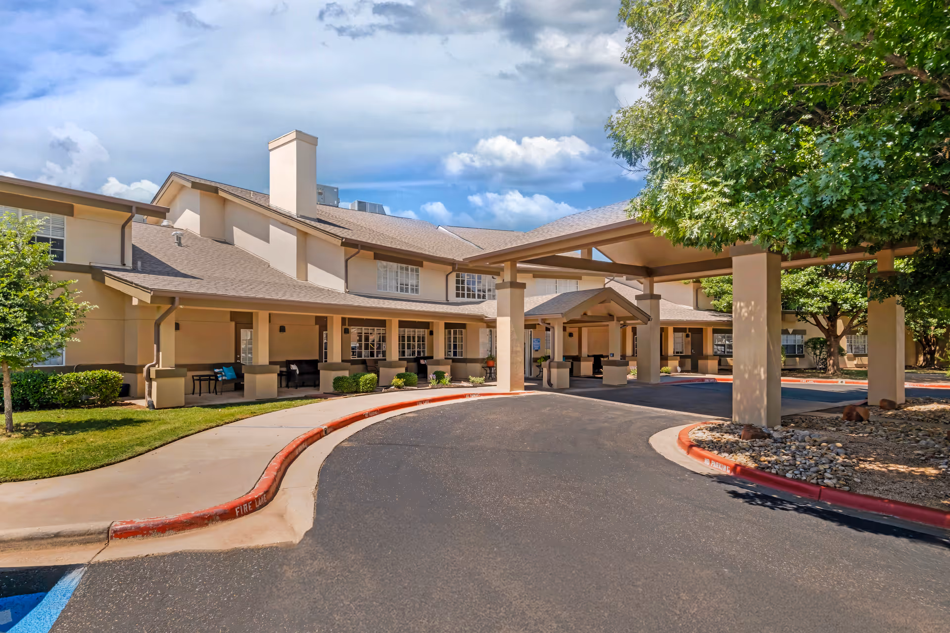 Exterior view of Brookdale Remington Park senior living facility showing a covered entrance with pillars, a curved driveway, landscaped greenery including trees and bushes, and a clear blue sky with some clouds.