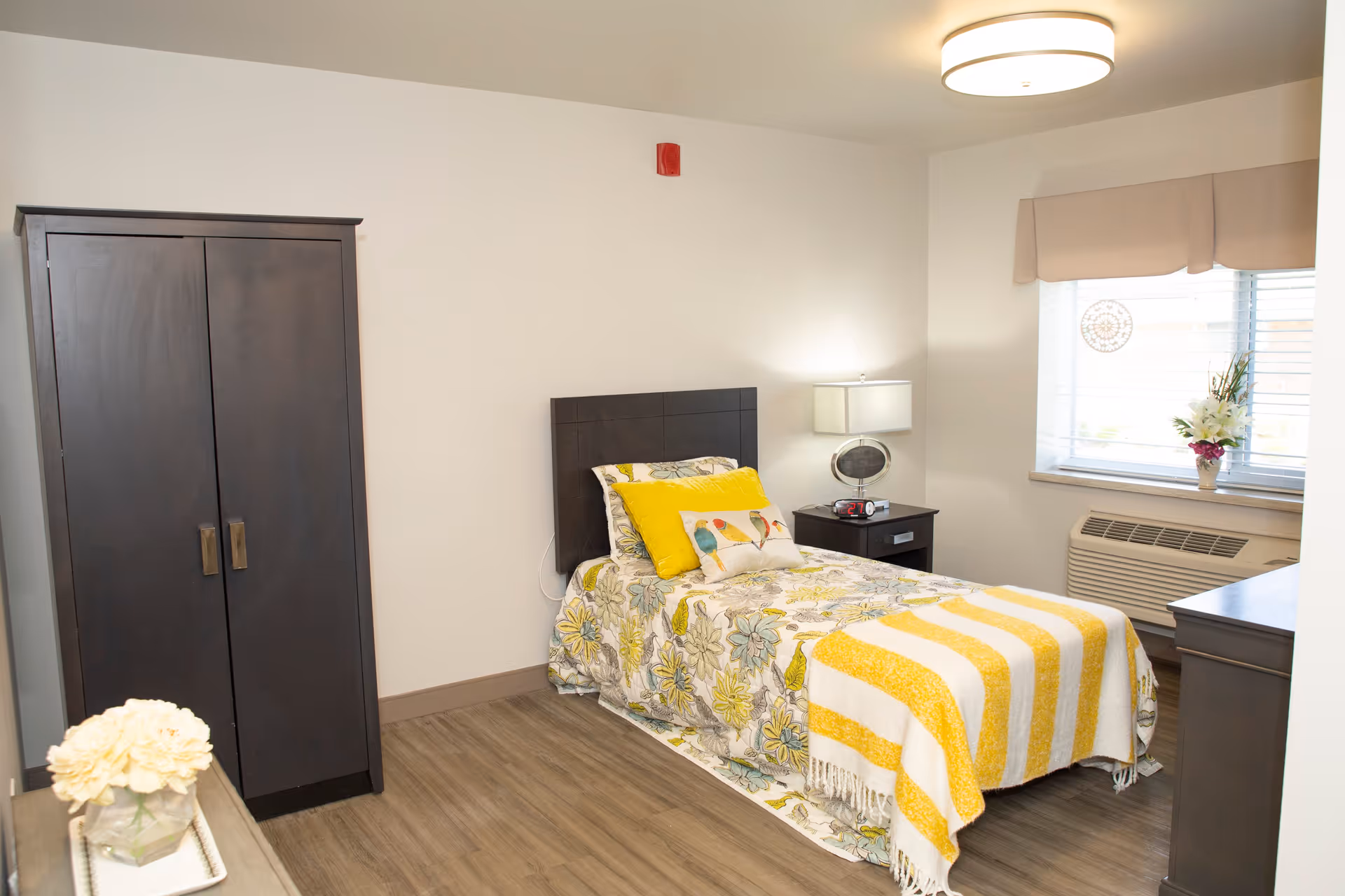A bright and clean bedroom with a single bed featuring floral and yellow bedding, a dark wooden wardrobe, a nightstand with a lamp and alarm clock, a window with blinds and a flower vase, and wood flooring.