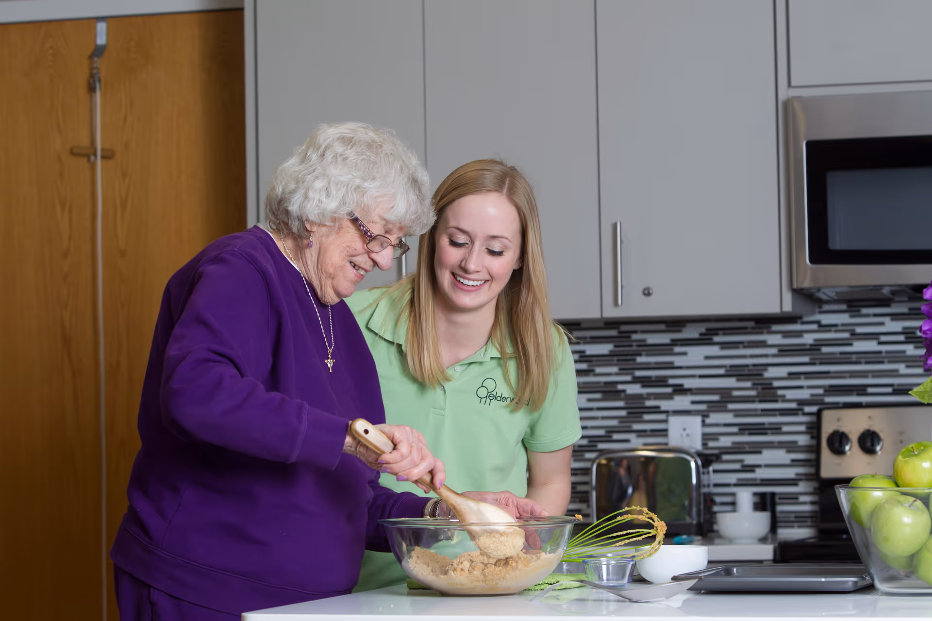 An elderly woman in a purple sweater is stirring a mixture in a glass bowl with a wooden spoon while a younger woman in a green Elderwood shirt smiles and watches her in a modern kitchen setting.