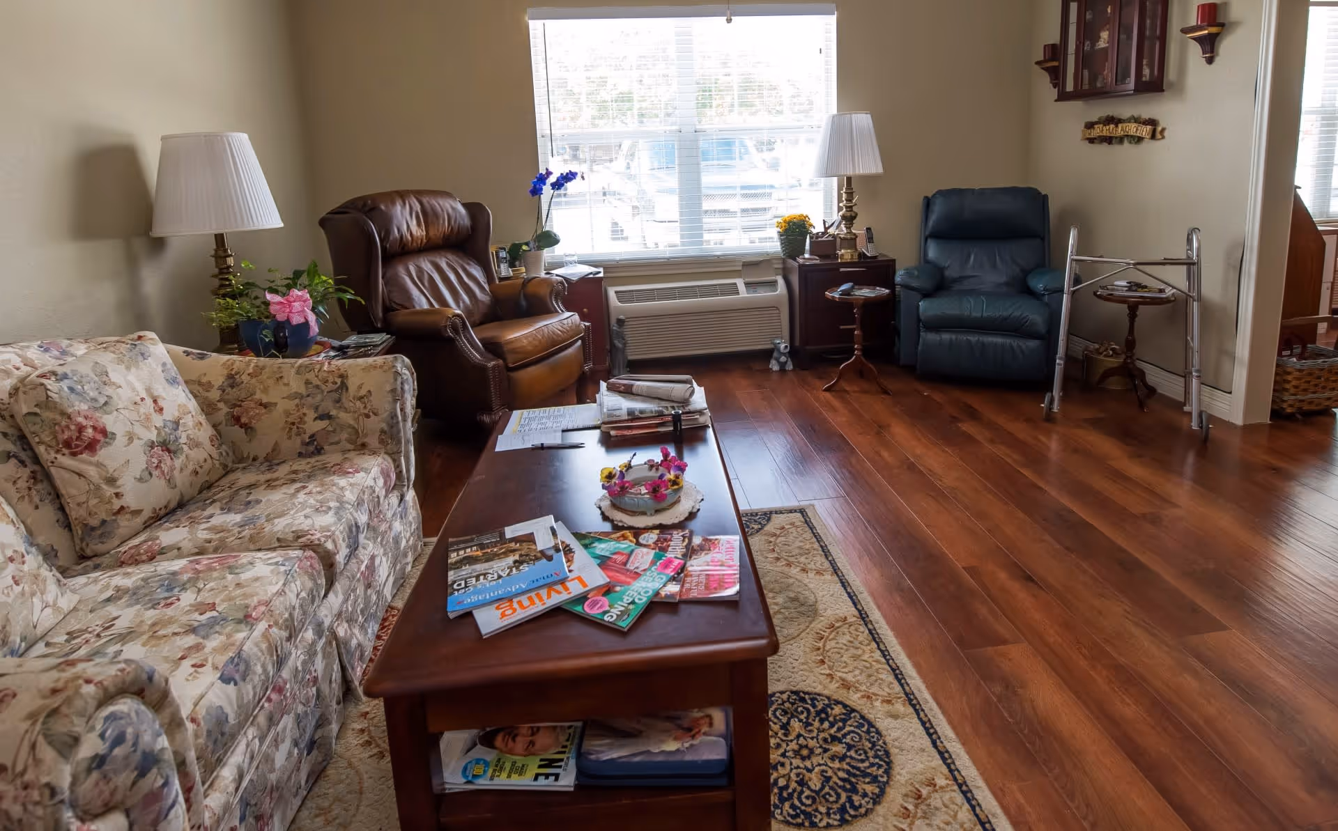 A cozy living room with a floral patterned sofa, a brown leather armchair, and a blue recliner chair. A wooden coffee table in the center holds magazines and a decorative flower arrangement. There are two side tables with lamps and plants, a window with blinds letting in natural light, and a walker near the blue recliner. The floor is wooden with a patterned area rug.