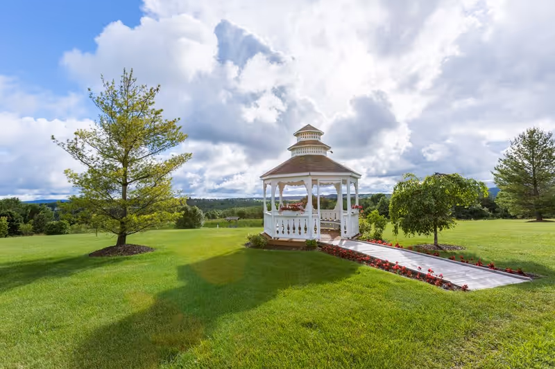White gazebo on a manicured green lawn with trees and a flower-lined walkway under a cloudy sky.