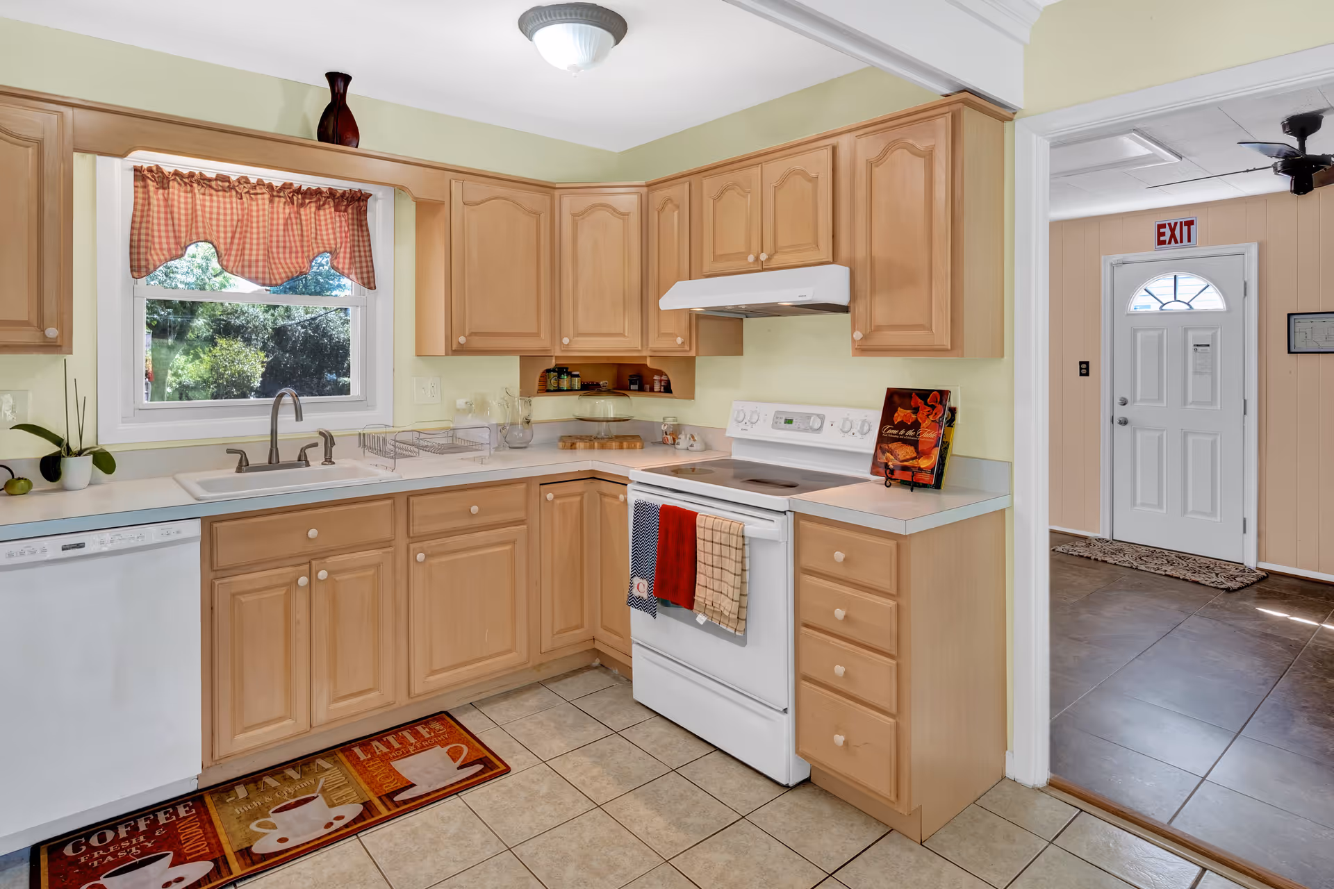 A bright kitchen with light wood cabinets, a white stove with three towels hanging on the handle, a white dishwasher, and a sink under a window with a red checkered valance. There is a colorful coffee-themed rug on the tiled floor and a white door with an exit sign visible in the adjacent room.
