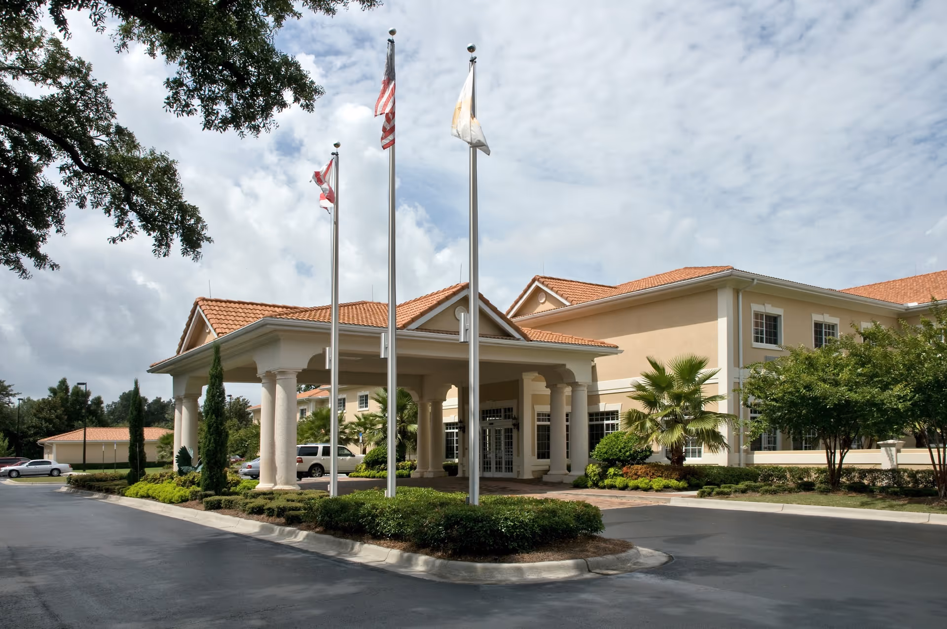 Exterior view of a senior living facility with a covered entrance supported by columns, three flagpoles with flags, landscaped greenery, and a clear sky with some clouds.