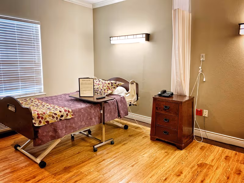 A rehabilitation center bedroom with a single hospital bed covered in a purple floral bedspread and a patterned pillow. There is a small overbed table with a framed document and a remote control on it. Next to the bed is a wooden nightstand with three drawers and a telephone on top. The room has wooden flooring, beige walls, a window with closed blinds, and a wall-mounted light fixture above the bed. A privacy curtain is partially visible on the right side.