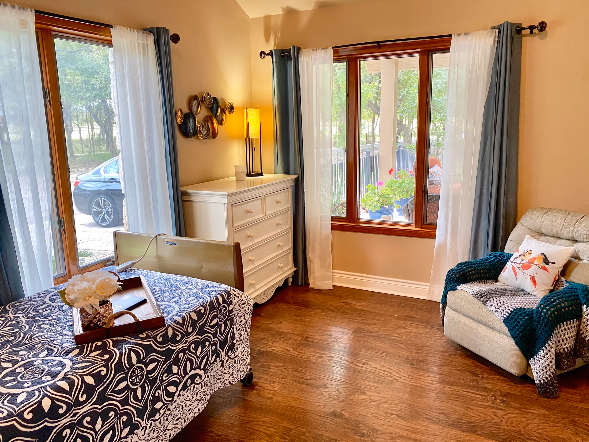 A cozy assisted living room with a hospital-style bed covered in a blue and white patterned blanket, a wooden tray with white flowers on the bed, a white dresser with a lamp and decorative wall art above it, large windows with sheer white and blue curtains letting in natural light, and a comfortable beige recliner chair with a bird-themed pillow and a knitted blanket.