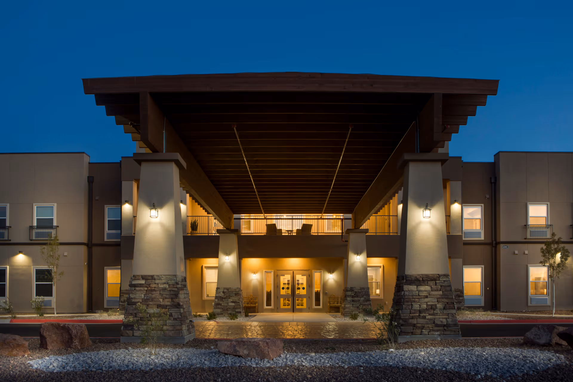 Exterior front view of The Legacy at Cimarron senior living facility at dusk, showing a large covered entrance with stone pillars and warm lighting illuminating the building.