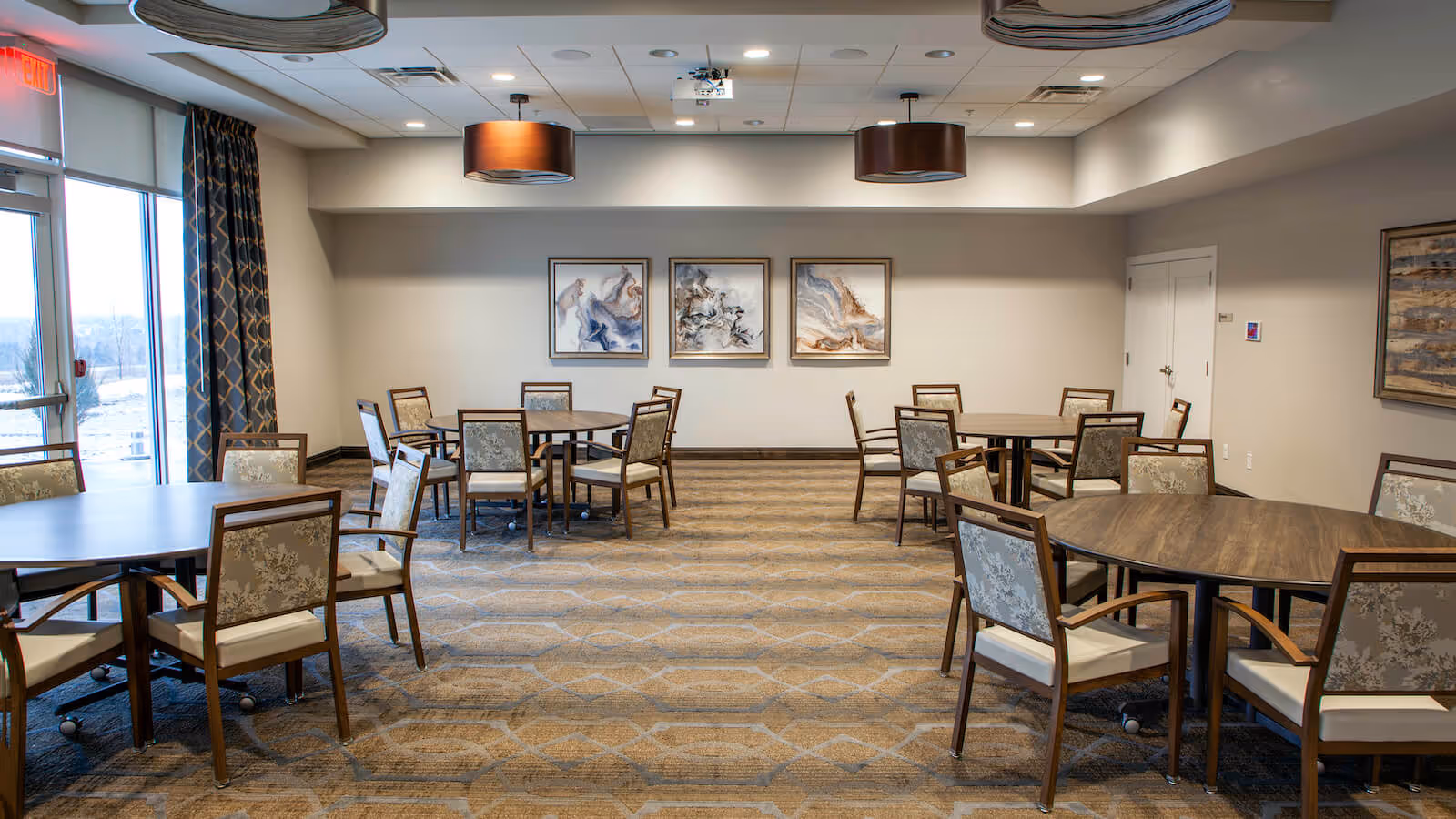 Communal dining/activity room with several round tables and chairs, wall art, pendant lights, and large windows.