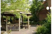 A wooden gazebo surrounded by green trees and bushes next to a brick building, with a paved walkway leading through the garden area.