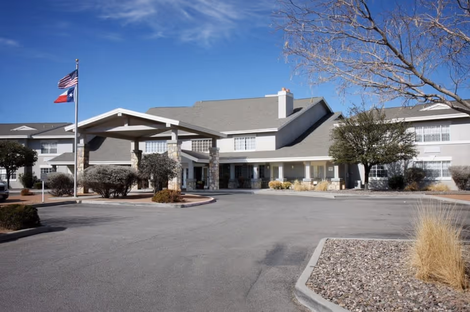 Front entrance of a two-story senior living facility with a covered porte-cochere, flagpoles, and landscaped parking area under a clear blue sky.