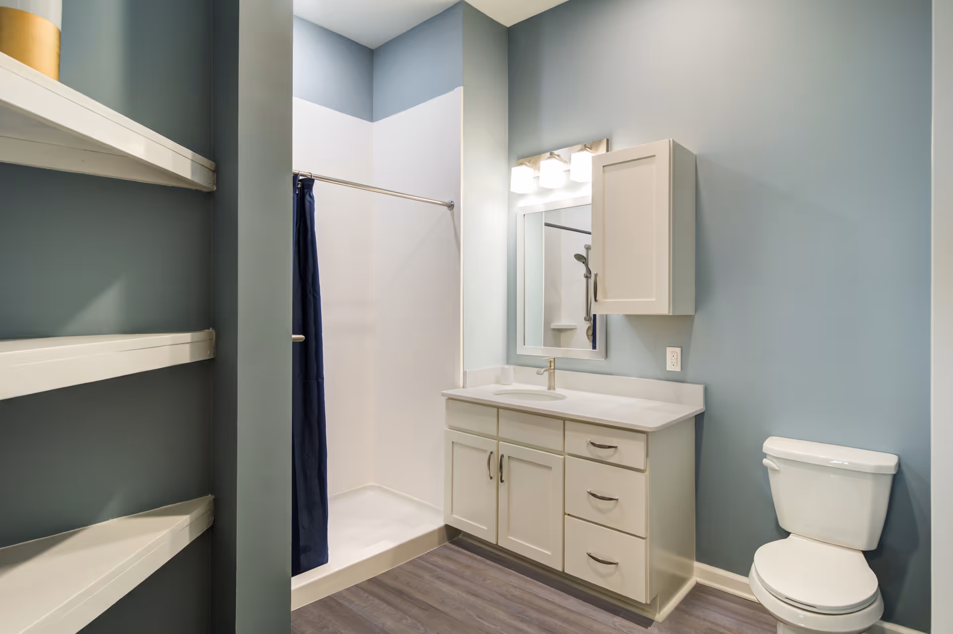 A clean bathroom with light blue walls featuring a white vanity with a sink and mirror above it, a white toilet, a shower with a dark blue curtain, and built-in white shelves on the left side.
