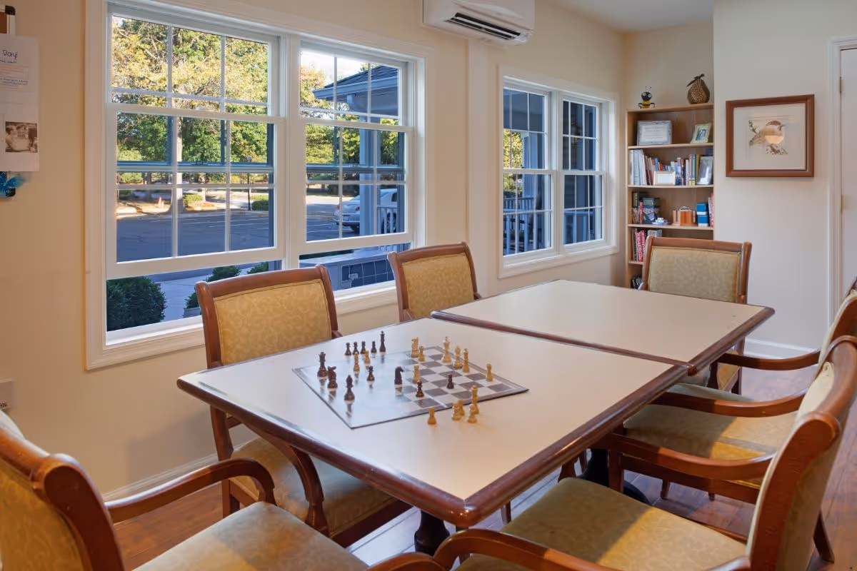 A well-lit room with a rectangular table surrounded by six cushioned wooden chairs. A chessboard with pieces set up is placed on the table. The room has large windows showing an outdoor parking area and greenery. A bookshelf filled with books and decorative items is against the wall, along with a framed picture and an air conditioning unit above the windows.